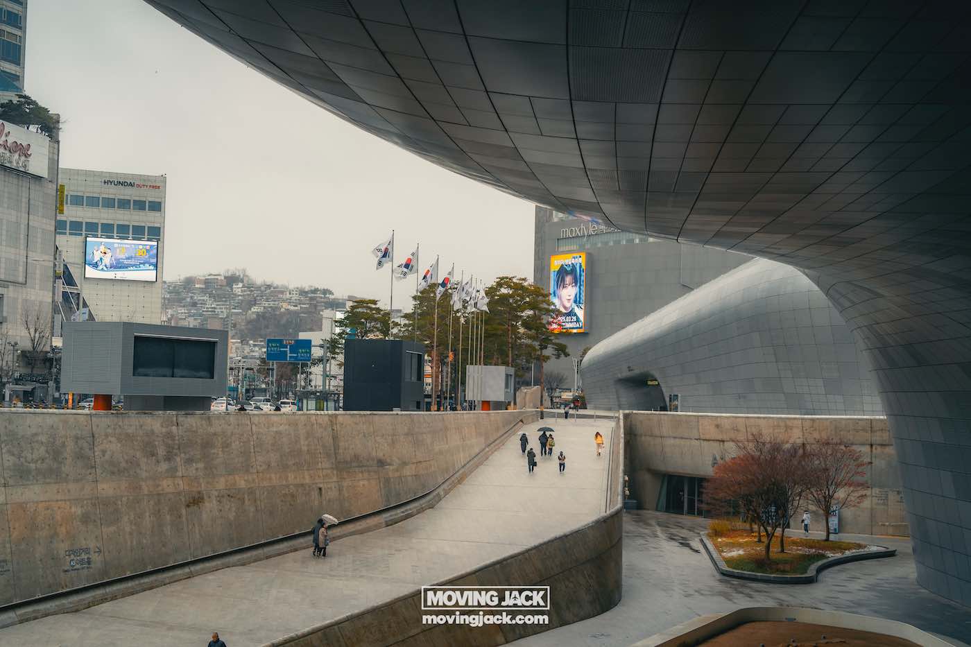 Wat te doen in Seoul: 13 unieke dingen om te doen + wat je beter kunt overslaan! 40 dingen om te doen in Seoul Moderne architectonische structuur met een gebogen ontwerp, groot open plein, vlaggen en wandelende mensen. Billboard met een kleurrijke advertentie. Bomen en stadsgezicht op de achtergrond. -copyright-moving-jack. Com