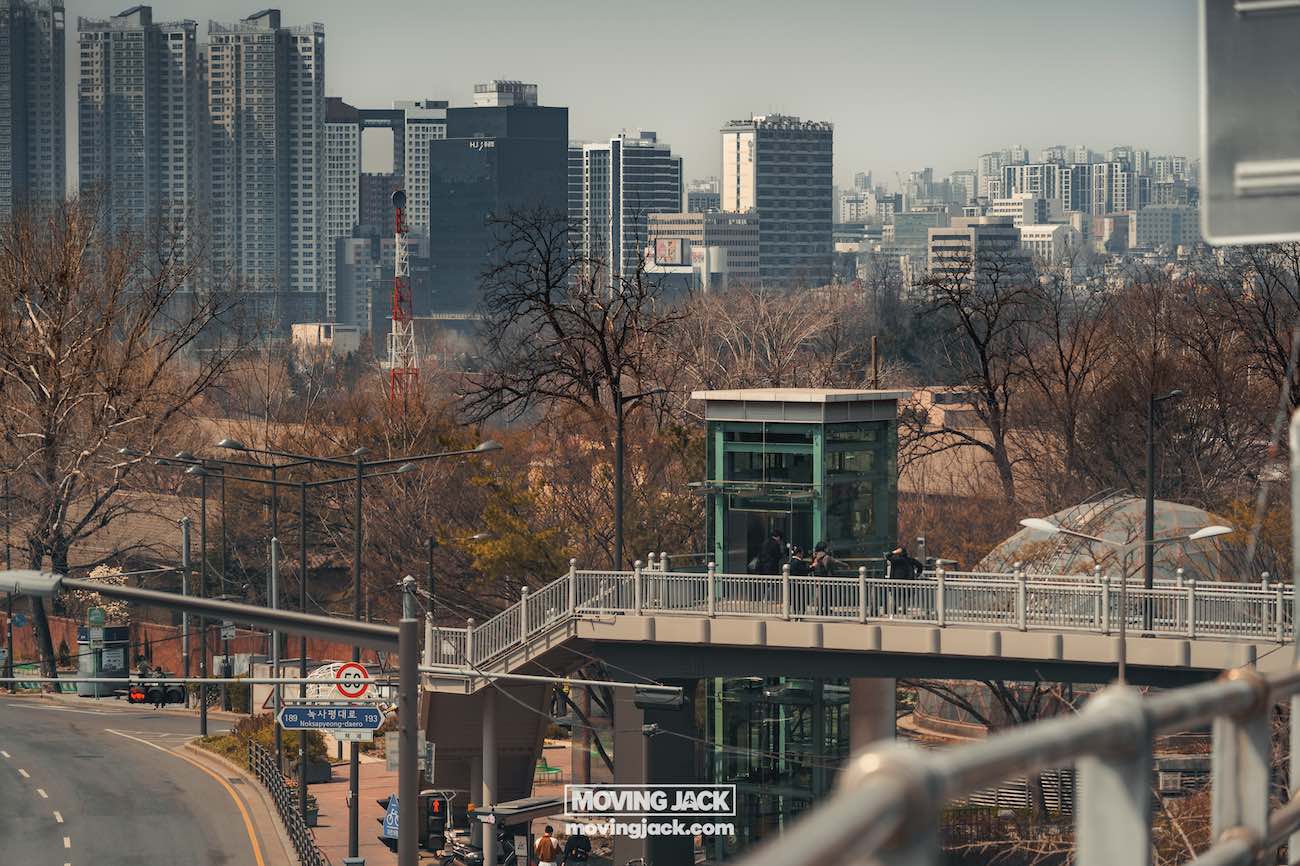Reisroute Itaewon 2025: de beste eetgelegenheden, drankjes en uitgaansgelegenheden 33 Itaewon Stedelijk landschap met een voetgangersbrug en lift, omgeven door kale bomen. Wolkenkrabbers en woongebouwen zijn zichtbaar op de achtergrond onder een bewolkte hemel. -copyright-moving-jack.com