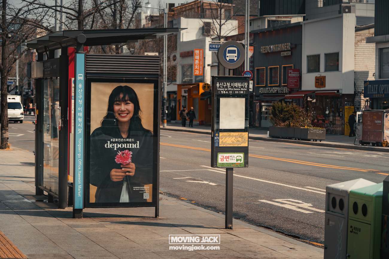 Reisroute Itaewon 2025: de beste eetgelegenheden, drankjes en uitgaansgelegenheden 34 itaewon Bushalte met een reclame waarop een vrouw bloemen vasthoudt, gelegen in een stadsstraat met winkels op de achtergrond. -copyright-moving-jack.com