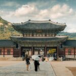 Seoul on a budget: save money in south korea 6 ikseon dong People walking toward the entrance of a traditional asian palace with mountains and cloudy sky in the background. -copyright-moving-jack. Com