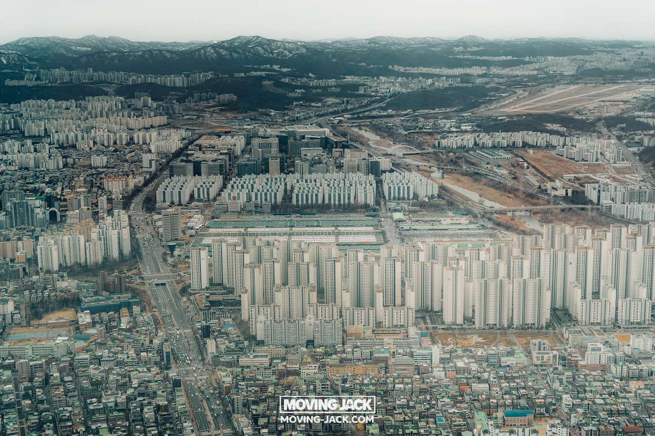 Luchtfoto van een dichtbevolkte stad met veel hoge flatgebouwen, wegen en groene ruimtes, omringd door bergen onder een bewolkte hemel – perfect om Seoul met een beperkt budget te verkennen. De tekst "moving jack" staat onderaan. -copyright-moving-jack.com