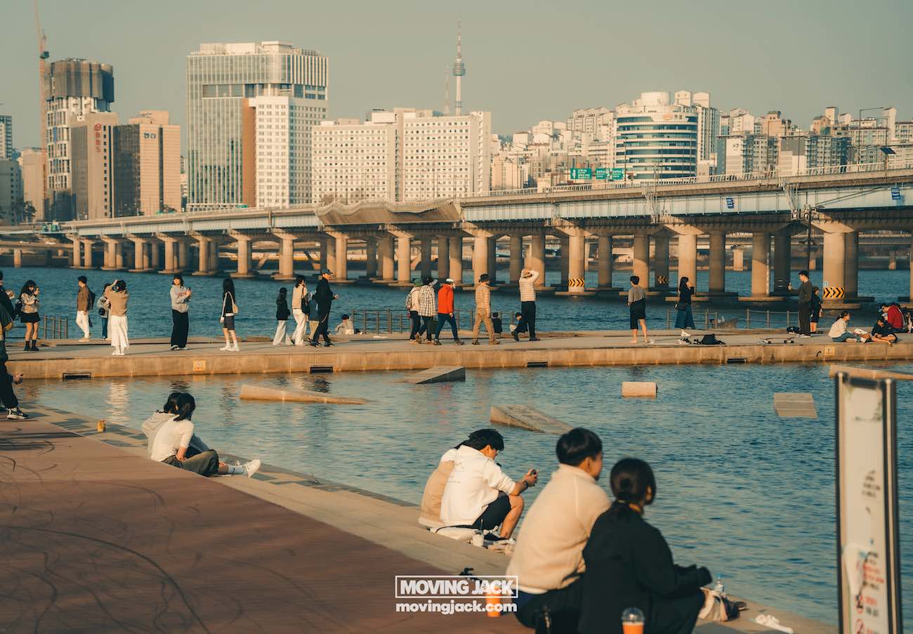 Seoul on a budget: save money in south korea 3 seoul on a budget People sitting and walking along the waterfront near a city skyline and bridge, with a clear sky in the background. -copyright-moving-jack. Com