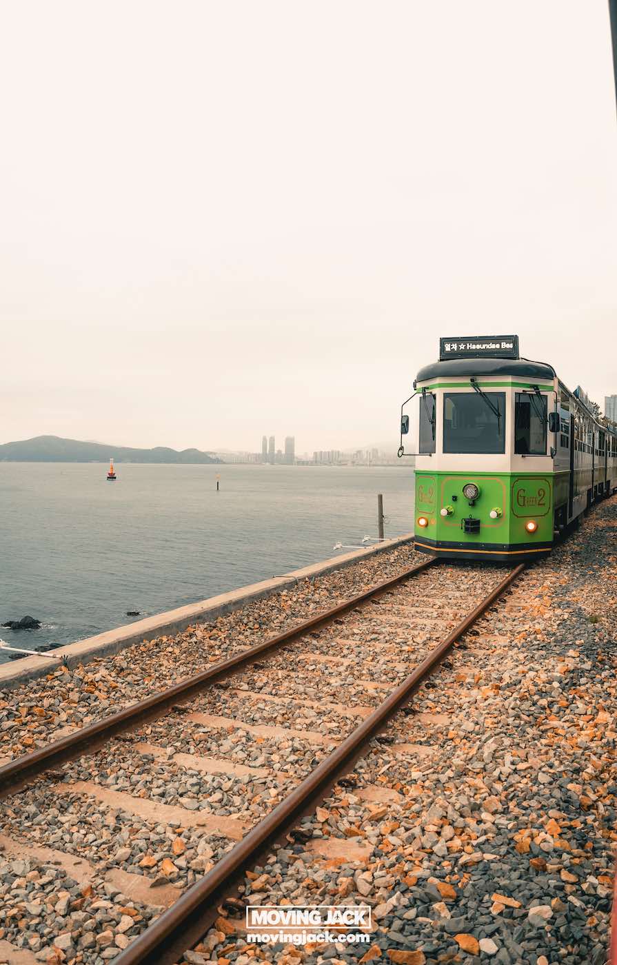 Gemakkelijk weekend in Busan met een peuter: 2-daagse reisroute voor gezinnen 20 Busan,weekend in Busan Een groene tram rijdt over rails langs een rotsachtige kustlijn, met stadsgebouwen zichtbaar in de verte onder een bewolkte hemel. -copyright-moving-jack.com