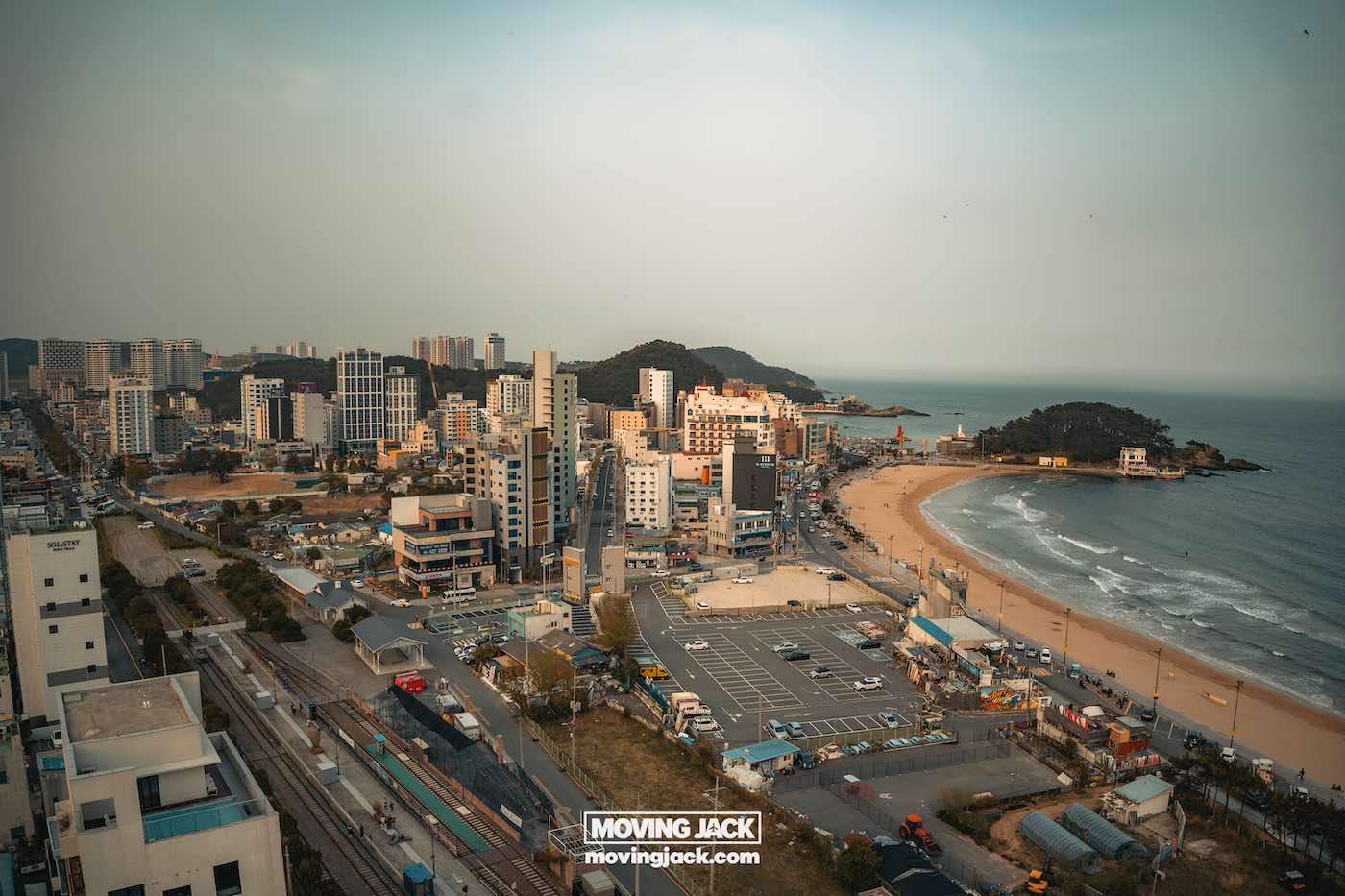Gemakkelijk weekend in Busan met een peuter: 2-daagse reisroute voor gezinnen 5 Busan,weekend in Busan Een kuststadsgezicht met hoge gebouwen, een zandstrand, spoorlijnen en een klein bebost eilandje vlak bij de kustlijn onder een heldere hemel. -copyright-moving-jack.com