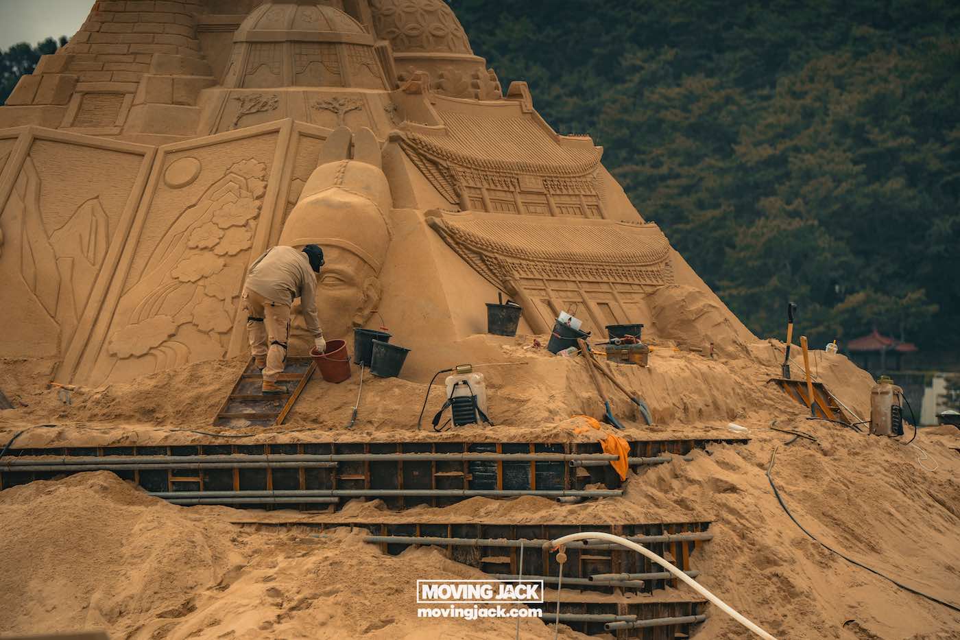 Gemakkelijk weekend in Busan met een peuter: 2-daagse reisroute voor gezinnen 23 Busan,weekend in Busan Een persoon werkt aan een groot, gedetailleerd zandsculptuur met architectonische elementen en een menselijk gezicht, omringd door gereedschap en emmers. -copyright-moving-jack.com