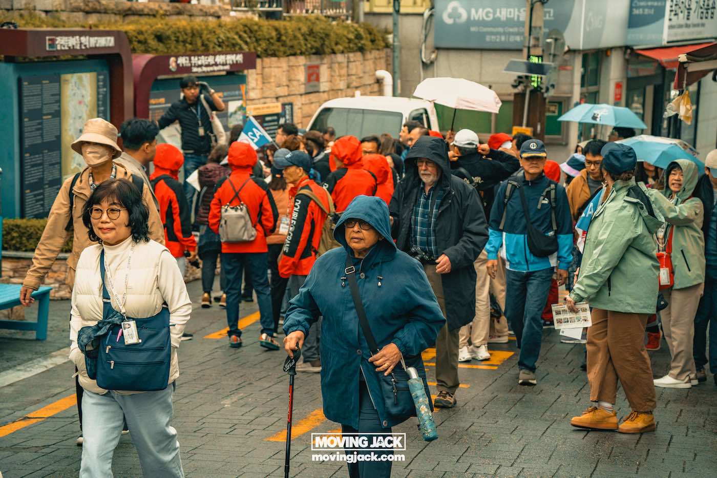 Gemakkelijk weekend in Busan met een peuter: 2-daagse reisroute voor gezinnen van 17 jaar en ouder in Busan, weekend in Busan Een groep mensen, velen met jassen en hoeden, loopt door een natte straat in een stedelijk gebied; sommigen dragen paraplu's en kaarten, wat een rondleiding door de stad op een regenachtige dag suggereert. -copyright-moving-jack.com