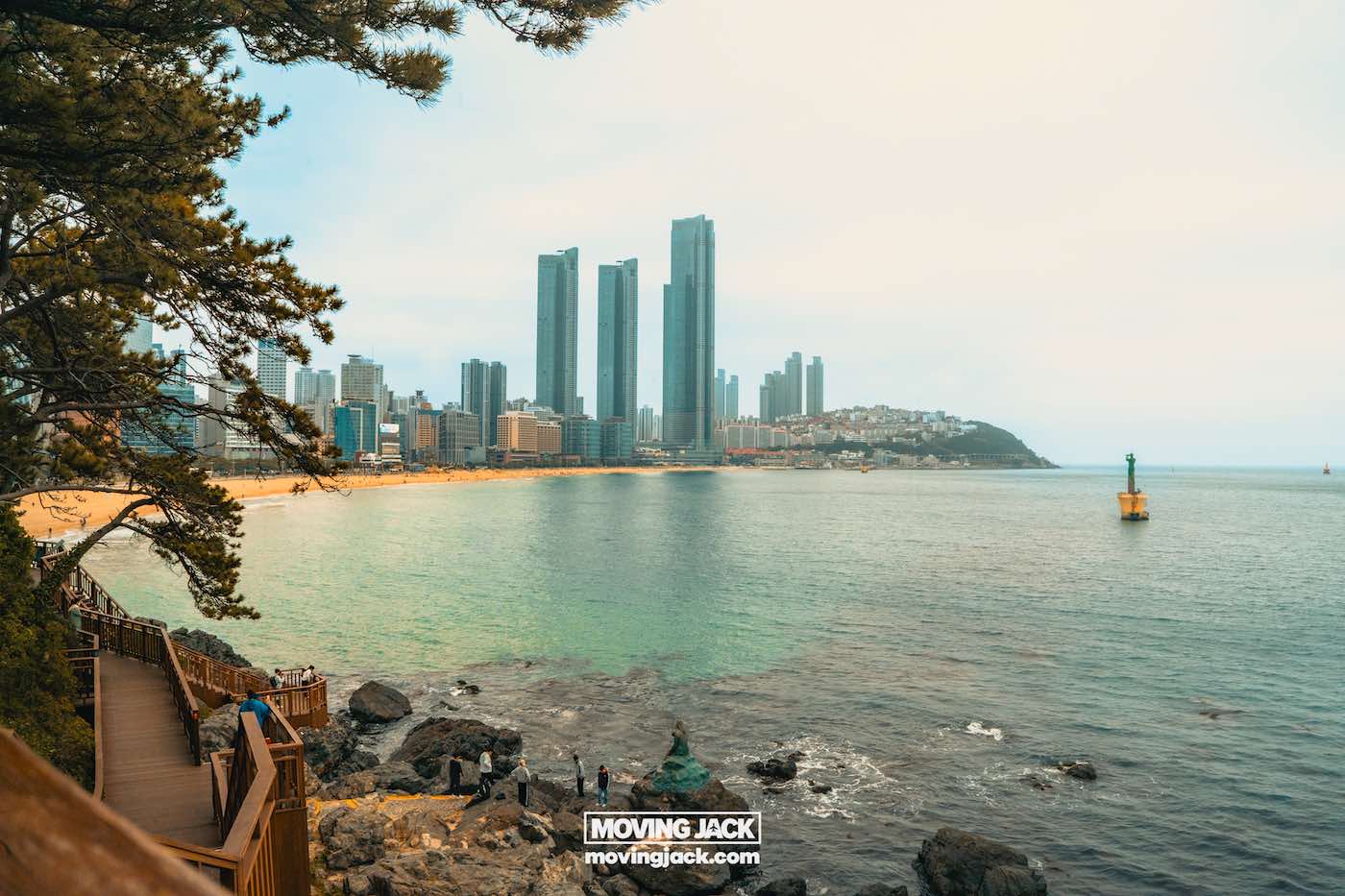 Gemakkelijk weekend in Busan met een peuter: 2-daagse reisroute voor gezinnen 21 Busan,weekend in Busan Uitzicht op een kuststad met hoge wolkenkrabbers, een zandstrand en een houten wandelpad langs de rotsachtige kustlijn; een groene vuurtoren staat in het water. -copyright-moving-jack.com