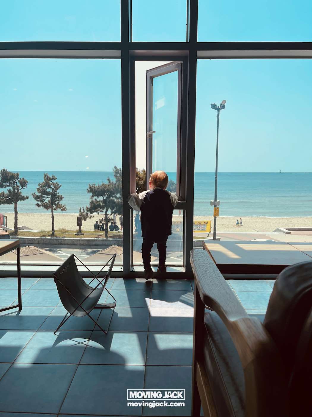 Gemakkelijk weekend in Busan met een peuter: 2-daagse reisroute voor gezinnen 3 Busan,weekend in Busan Een peuter staat in een open glazen deur en kijkt uit over een strand en de oceaan onder een helderblauwe lucht op een zonnige dag. -copyright-moving-jack.com
