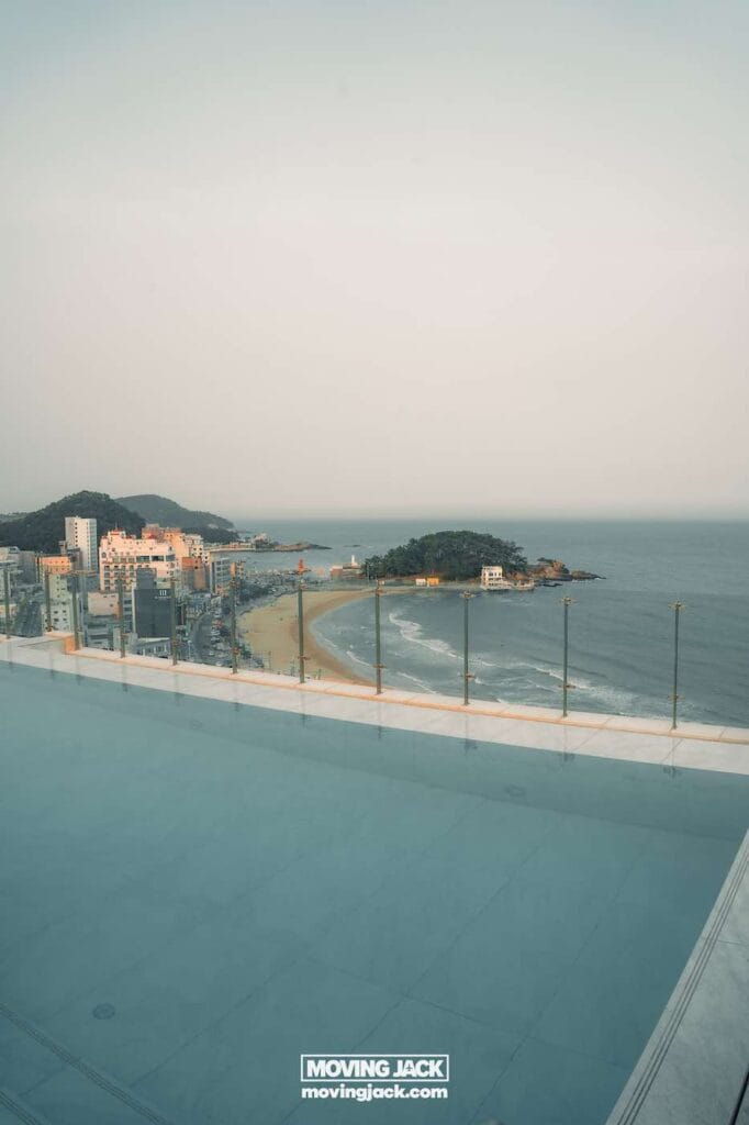 Gemakkelijk weekend in Busan met een peuter: 2-daagse reisroute voor gezinnen 6 Busan, weekend in Busan Uitzicht vanaf een zwembad op het dak over een kuststad, een zandstrand en een klein eiland, met gebouwen en heuvels op de achtergrond onder een wazige hemel. -copyright-moving-jack.com