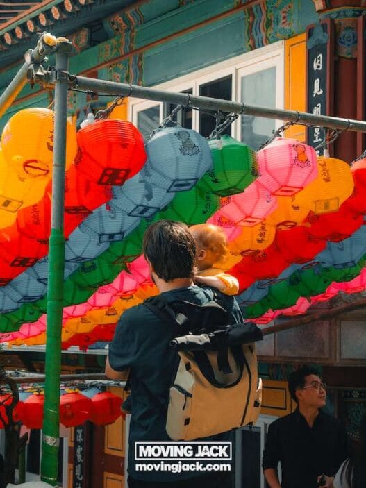 Easy weekend in busan with a toddler: 2-day itinerary for families 24 moving to south korea with a cat A man holding a child stands under a canopy of colorful paper lanterns outside a traditional building, with other people nearby. -copyright-moving-jack. Com