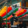 A man holding a child stands under a canopy of colorful paper lanterns outside a traditional building, with other people nearby. -copyright-moving-jack. Com