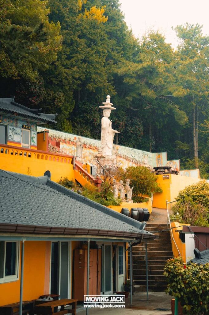 Gemakkelijk weekend in Busan met een peuter: 2-daagse reisroute voor gezinnen 15 Busan,weekend in Busan Een wit Boeddhabeeld staat op een verhoogd terras, omringd door gele gebouwen en groen, met trappen die naar het beeld leiden. -copyright-moving-jack.com