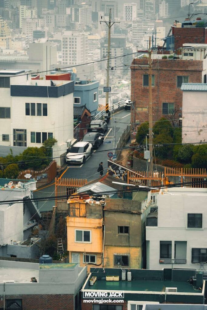 Gemakkelijk weekend in Busan met een peuter: 2-daagse reisroute voor gezinnen van 16 jaar en ouder in Busan, weekend in Busan Een stadsstraat met geparkeerde auto's, een persoon die op de stoep loopt en verschillende gebouwen, waaronder een geel gebouw op de voorgrond, onder een bewolkte hemel. -copyright-moving-jack.com