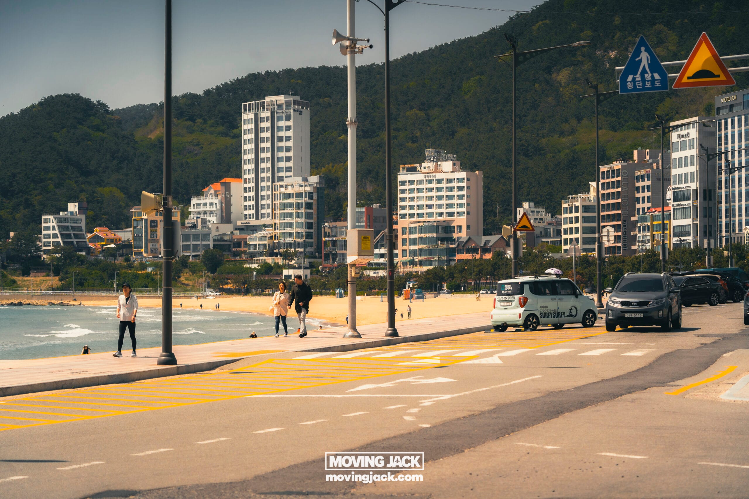 Gemakkelijk weekend in Busan met een peuter: 2-daagse reisroute voor gezinnen 30 Busan,weekend in Busan Een kuststraat met geparkeerde auto's langs de weg, mensen die op de stoep lopen en moderne gebouwen op de achtergrond vlakbij het strand. -copyright-moving-jack.com