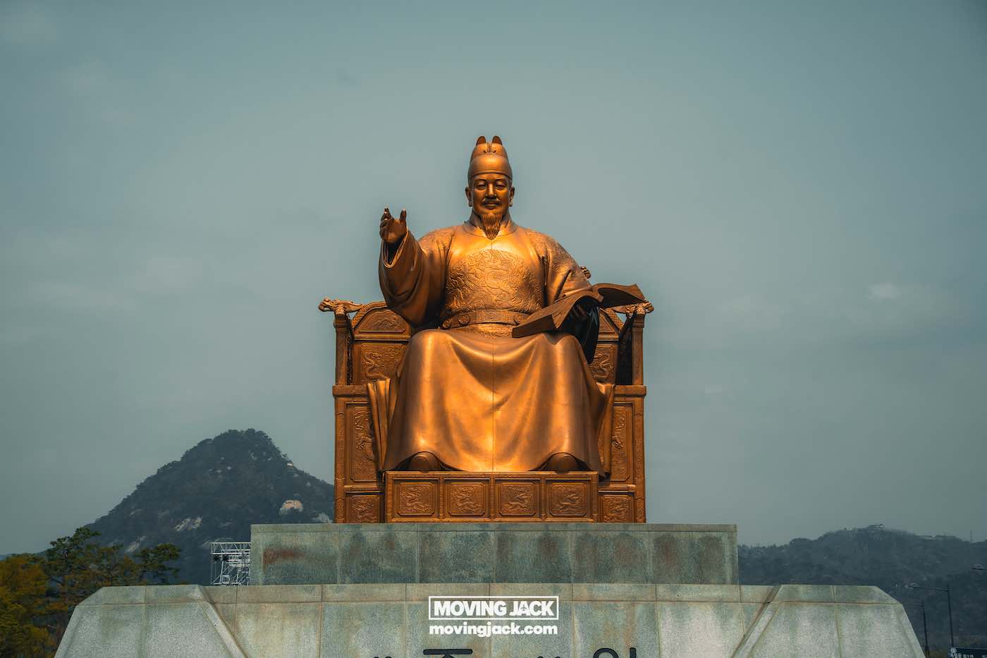 Gyeongbokgung paleis Seoul ultieme gids + wandelvideo 21 Gyeongbokgung Bronzen beeld van een zittende figuur in traditionele Koreaanse kledij, met een boek in de hand, tegen de achtergrond van bergen en de blauwe lucht nabij Gyeongbokgung. -copyright-moving-jack.com