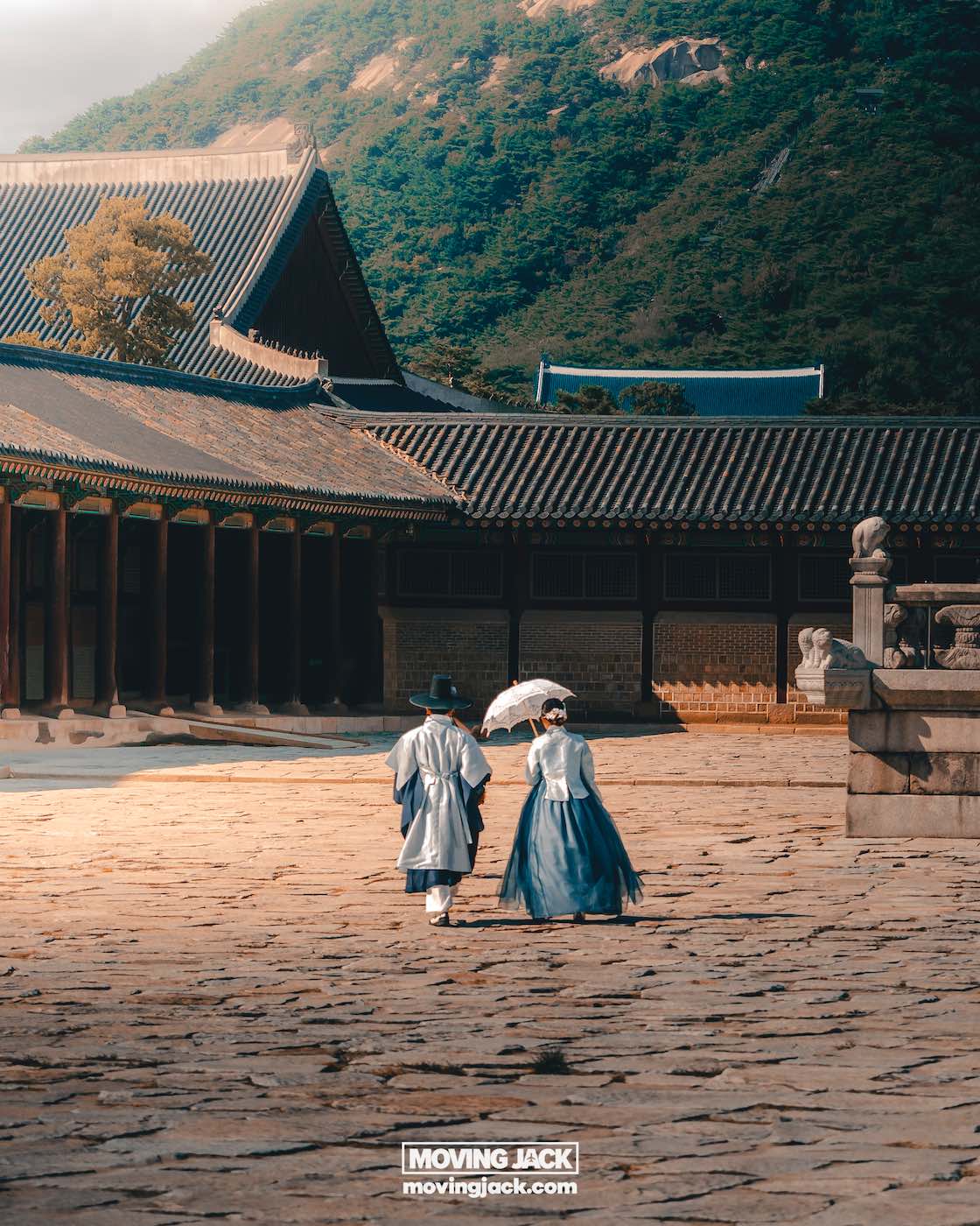 Gyeongbokgung paleis Seoul ultieme gids + wandelvideo 17 Gyeongbokgung Twee mensen in traditionele Koreaanse kleding lopen samen op de binnenplaats van het historische Gyeongbokgung-paleis, met betegelde daken en een met bomen bedekte heuvel op de achtergrond. -copyright-moving-jack.com