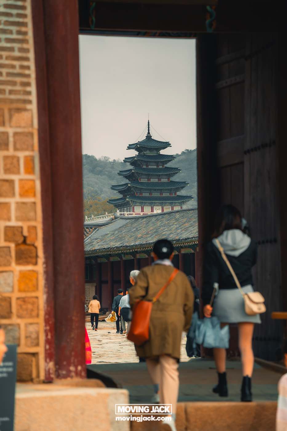 Gyeongbokgung paleis Seoul ultieme gids + wandelvideo 26 Gyeongbokgung Mensen lopen door een poort naar een traditionele pagode met meerdere verdiepingen op de historische Gyeongbokgung-locatie, met bergen zichtbaar op de achtergrond. -copyright-moving-jack.com