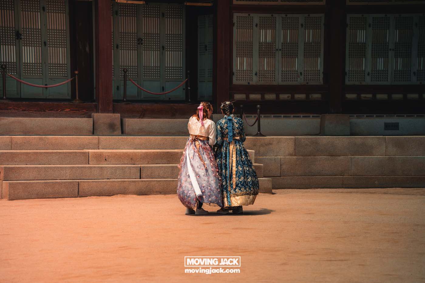 Gyeongbokgung paleis Seoul ultieme gids + wandelvideo 10 Gyeongbokgung Twee mensen in traditionele Koreaanse hanbok lopen zij aan zij voor stenen trappen en een houten gebouw in Gyeongbokgung. -copyright-moving-jack.com