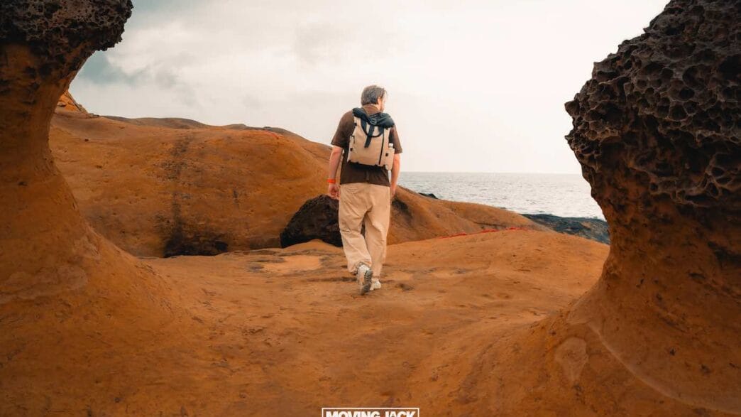 Een persoon met een rugzak, geïnspireerd door reiscitaten, loopt over oranje, rotsachtig terrein richting de oceaan onder een bewolkte hemel. -copyright-moving-jack.com