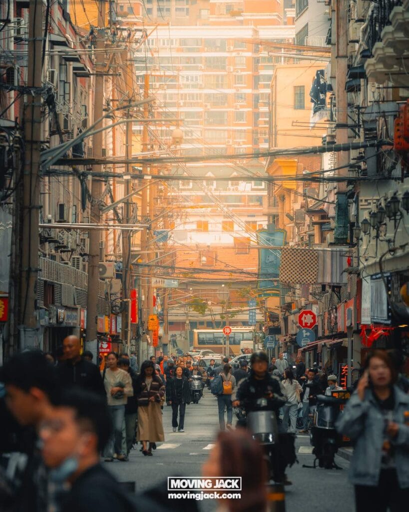 Busy urban street scene with people walking and riding scooters, flanked by tall buildings and dense overhead wires under soft, warm light—capturing the vibrant atmosphere for those exploring where to stay in Shanghai. -Copyright-moving-jack.com