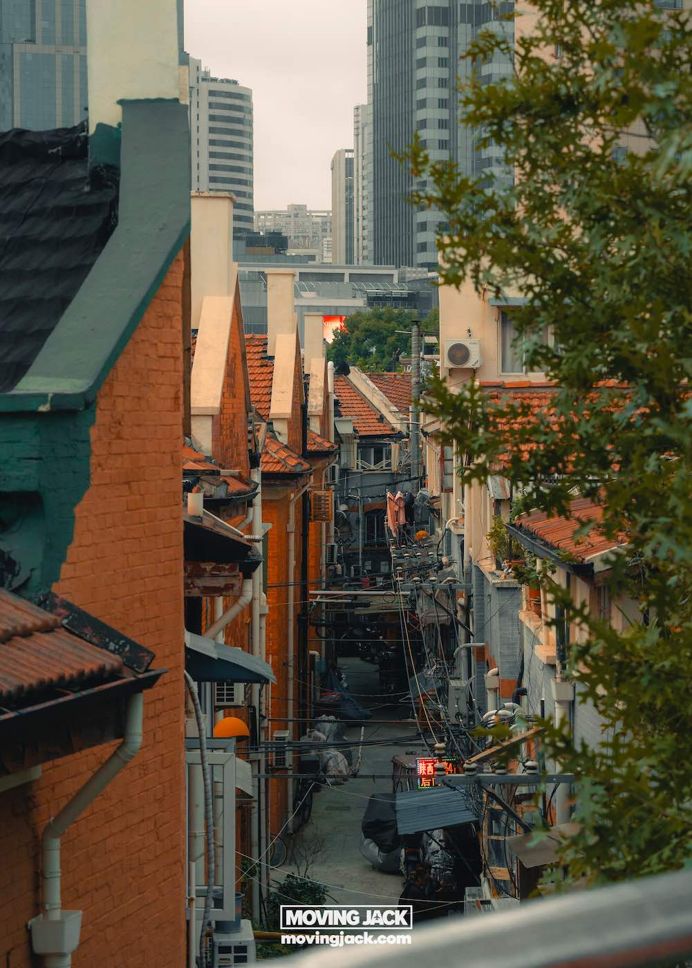 Waar te verblijven in Shanghai: 5 leukste wijken + beste hotels 13 waar te verblijven in Shanghai Smal steegje met oude gebouwen en een wirwar van draden, met moderne wolkenkrabbers op de achtergrond onder een bewolkte hemel; "Moving Jack"-borden onderaan – een authentieke blik op waar je in Shanghai kunt overnachten. -copyright-moving-jack.com