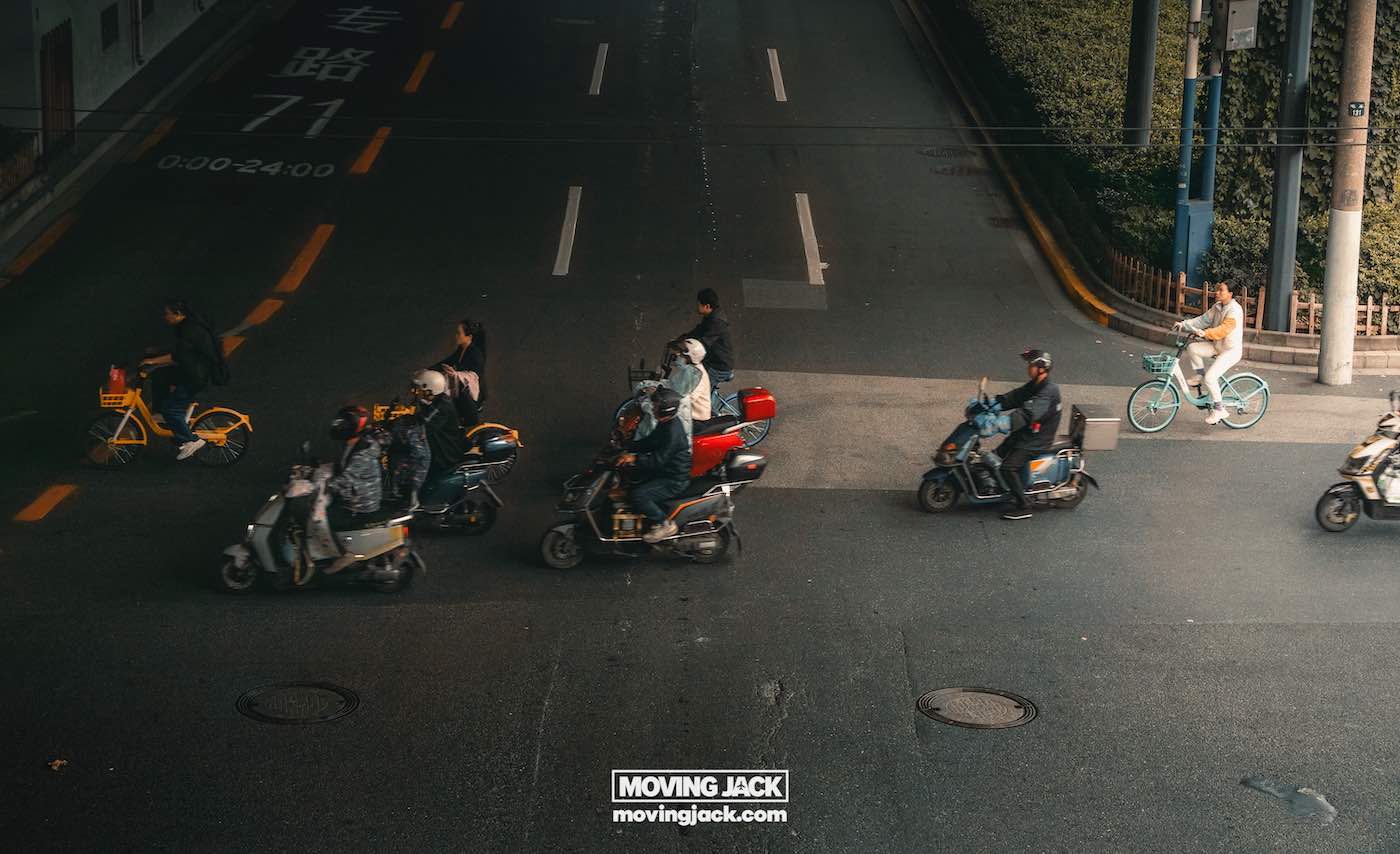 Waar te verblijven in Shanghai: 5 leukste wijken + beste hotels 24 waar te verblijven in Shanghai Verschillende mensen rijden op scooters en fietsen door een stadsstraat, sommigen wachten bij een oversteekplaats, terwijl één fietser over een aparte rijstrook rijdt onder bovenverlichting – een treffende weergave van de levendige energie van het verkennen van de beste plekken om te overnachten in Shanghai. -copyright-moving-jack.com