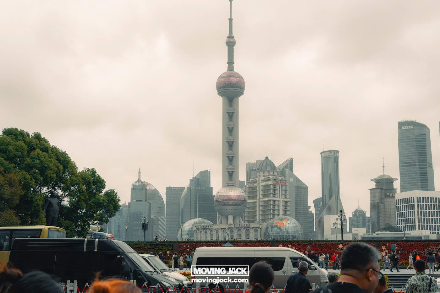 Waar te verblijven in Shanghai: 5 leukste wijken + beste hotels 3 waar te verblijven in Shanghai Een blik op de skyline van Shanghai met de Oriental Pearl Tower op een bewolkte dag, met mensen en voertuigen op de voorgrond – een must-see voor iedereen die zich afvraagt waar te overnachten in Shanghai. -copyright-moving-jack.com