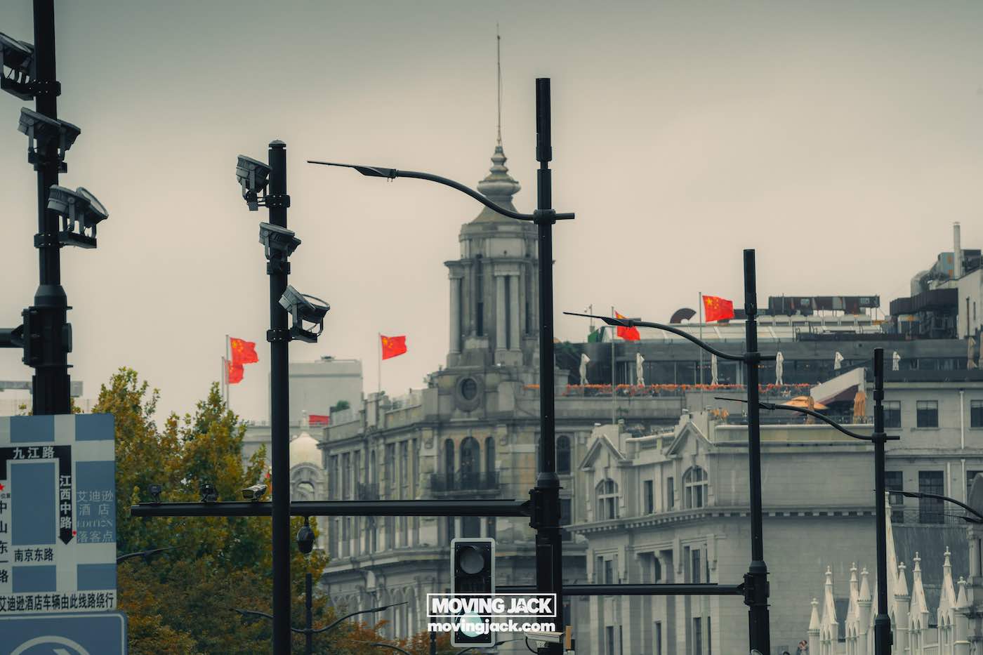 Waar te verblijven in Shanghai: 5 leukste wijken + beste hotels 4 waar te verblijven in Shanghai Stedelijk tafereel met meerdere bewakingscamera's, Chinese vlaggen en historische gebouwen onder een bewolkte hemel. Onderaan staat een bord met de tekst "moving jack movingjack.com" – een glimp van het stadsleven voor wie zich afvraagt waar te overnachten in Shanghai. -copyright-moving-jack.com