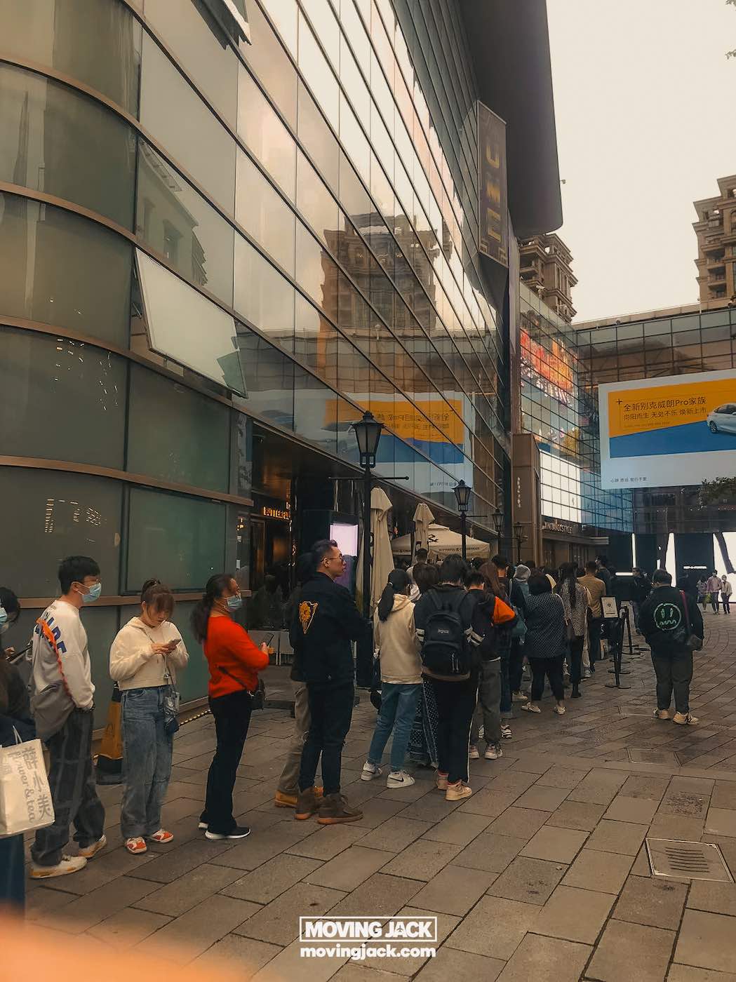 Waar te verblijven in Shanghai: 5 leukste wijken + beste hotels 28 waar te verblijven in Shanghai Mensen staan in de rij voor een modern glazen gebouw op een bewolkte dag; de meesten dragen jassen en mondkapjes, misschien wachtend op aanwijzingen over waar ze in Shanghai kunnen overnachten. -copyright-moving-jack.com