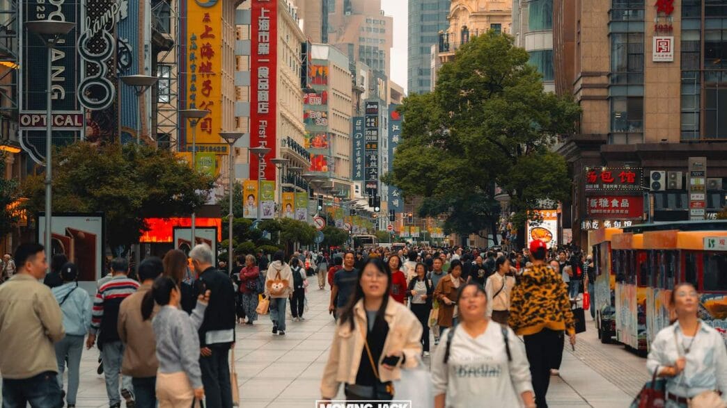 Een drukke stadsstraat in Shanghai met veel mensen die er lopen, hoge gebouwen met kleurrijke borden en bomen langs de wandelpaden – perfect om te verkennen terwijl je overdag beslist waar je in Shanghai wilt verblijven. -copyright-moving-jack.com