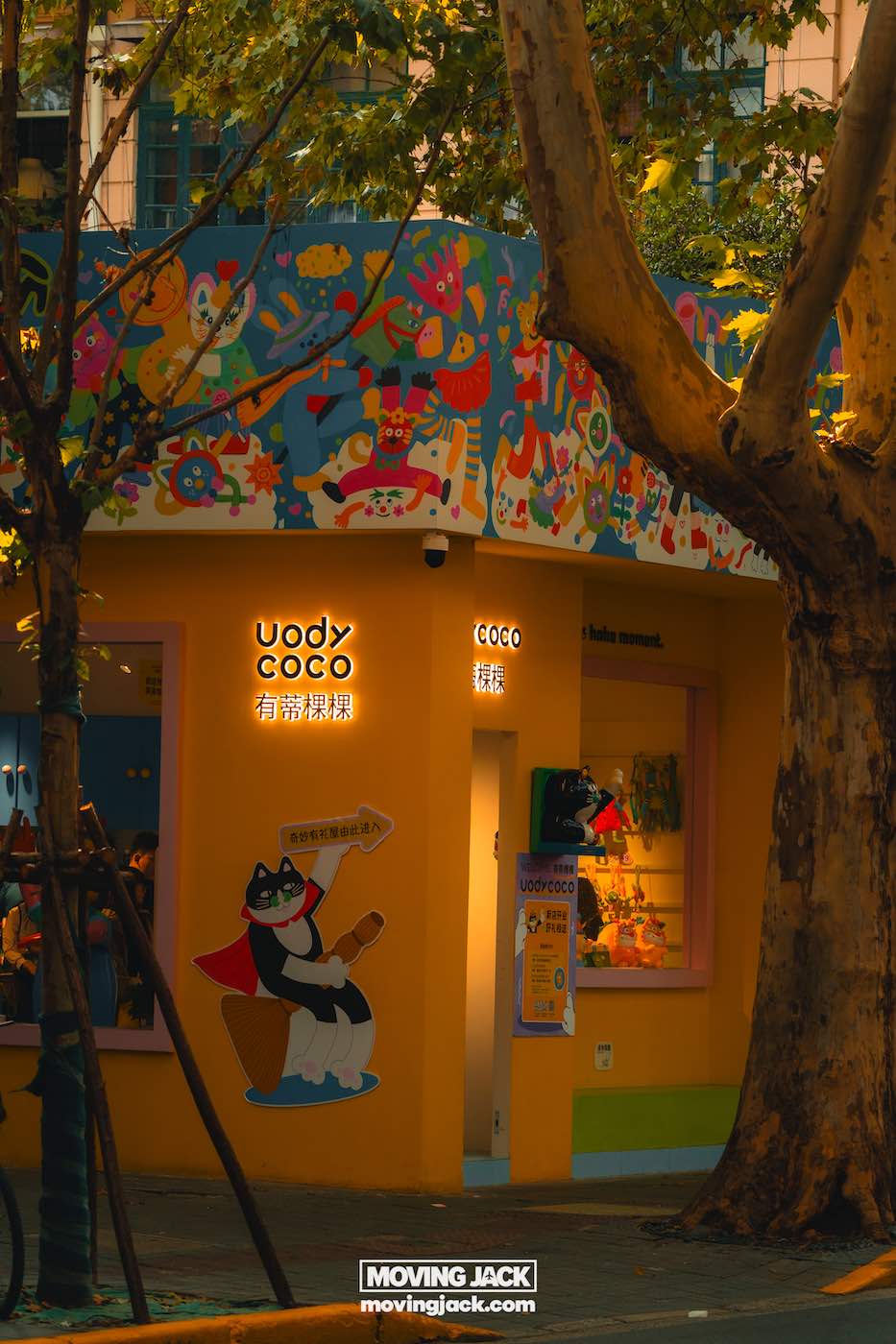 Waar te verblijven in Shanghai: 5 leukste wijken + beste hotels 15 waar te verblijven in Shanghai Felgekleurde winkelpui met "coco"-borden, een muurschildering met een cartoonkat en verlichte lampen, gedeeltelijk in de schaduw van grote bomen; een gezellig straatbeeld dat charme toevoegt aan wie op zoek is naar een verblijfplaats in Shanghai. -copyright-moving-jack.com