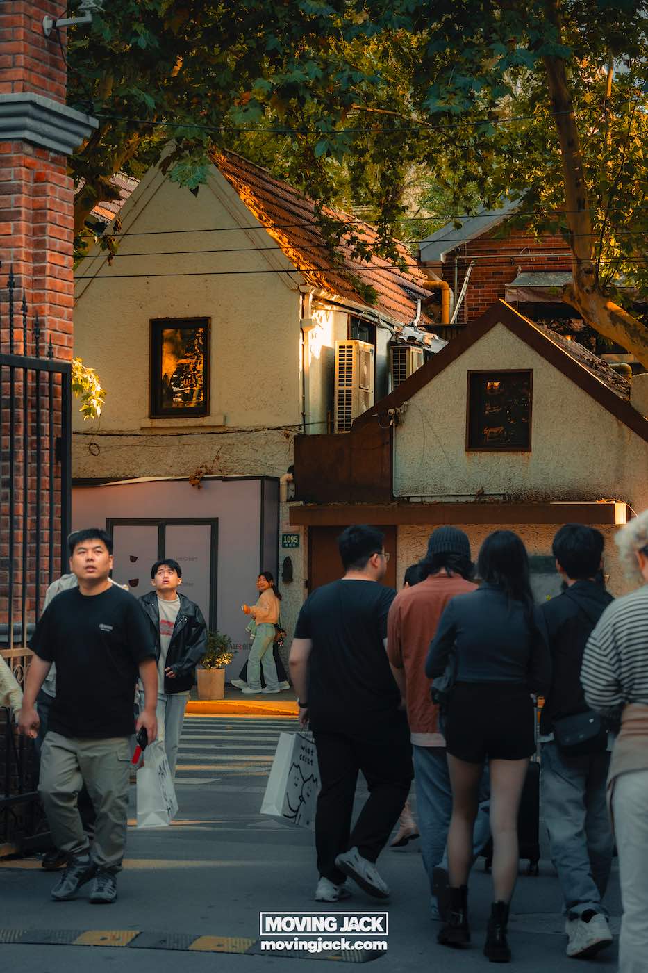 Waar te verblijven in Shanghai: 5 leukste wijken + beste hotels 12 waar te verblijven in Shanghai Een groep mensen loopt op een stadsstraat bij een poort, met een klein huis en bomen op de achtergrond. Op de voorgrond verschijnt een bord met het logo "moving jack", dat aangeeft waar je in Shanghai kunt overnachten. -copyright-moving-jack.com