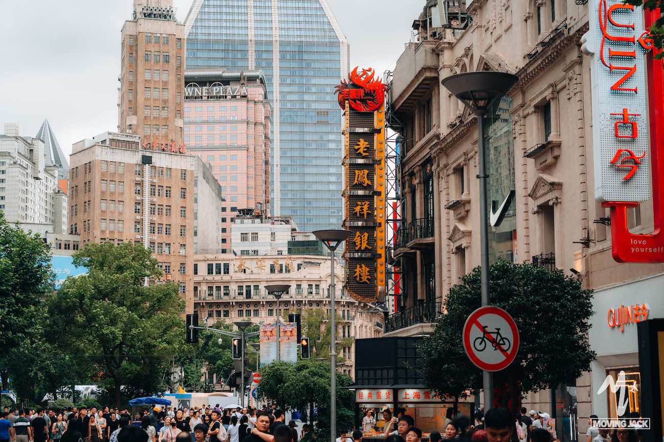 A bustling city street filled with people, lined with trees and tall buildings. Prominent Chinese signage hints at where to stay in Shanghai, with modern skyscrapers rising in the background under a cloudy sky. -Copyright-moving-jack.com