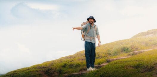 A man wearing outdoor gear carries a child in a backpack carrier and points ahead while standing on a grassy hill—an ideal moment for any travel blog or moving jack adventure. -copyright-moving-jack. Com