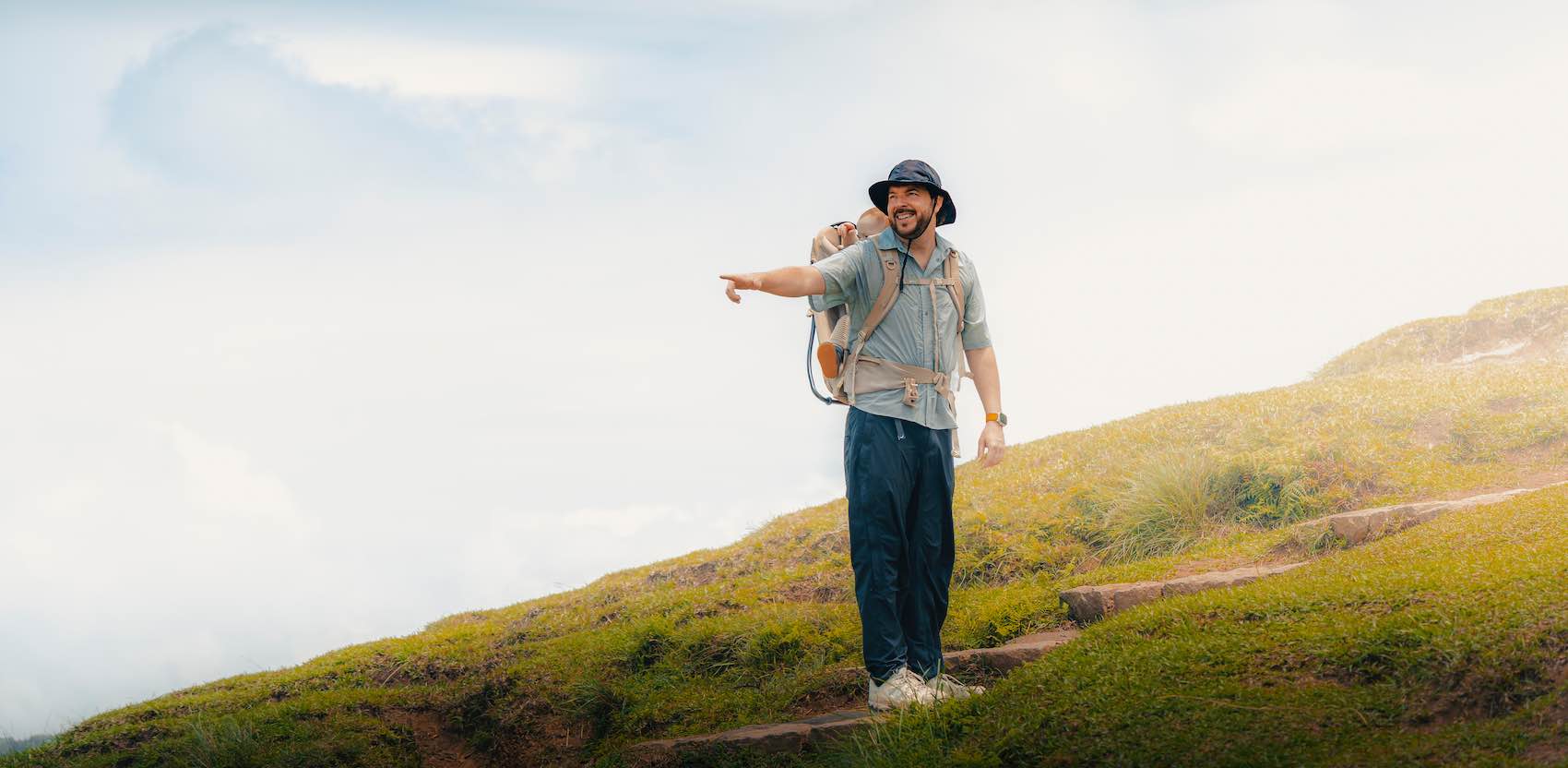 Verhuiskrik voor verhuiskrik, reisblog Een man in outdoorkleding draagt een kind in een rugzak en wijst vooruit terwijl hij op een grasheuvel staat – een ideaal moment voor een reisblog of een avontuur met een verhuiswagen. -copyright-moving-jack.com