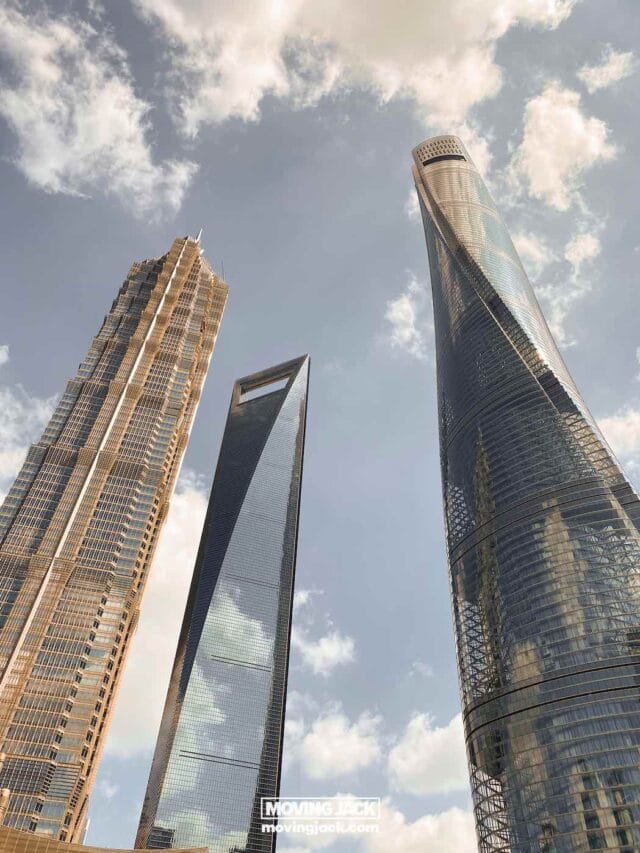 Drie moderne wolkenkrabbers met opvallende ontwerpen steken af tegen een gedeeltelijk bewolkte hemel, gezien vanaf de grond. De gebouwen zijn voorzien van reflecterend glas en unieke vormen, waarmee ze hedendaagse stedelijke architectuur tentoonstellen. -Copyright-moving-jack.com