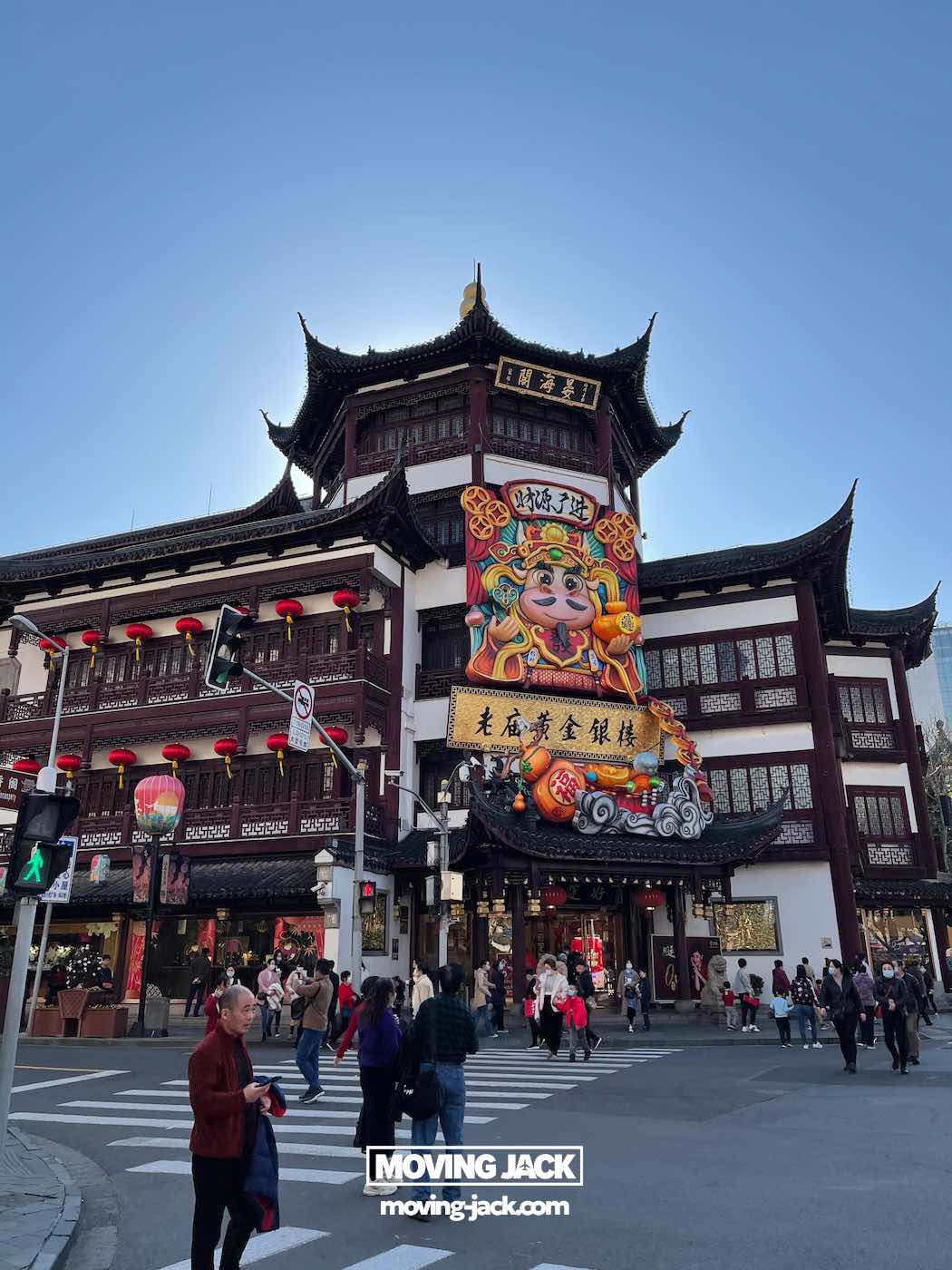 Is je tuin een bezoek waard in 2025? Complete gids & eerlijke lokale review 8 Is je tuin een bezoek waard? Voetgangers steken de straat over voor een traditioneel Chinees gebouw vlakbij Yu Garden, versierd met lantaarns en een feestelijke banner met een tekenfilmfiguur, die laat zien waarom Yu Garden een bezoek waard is. -copyright-moving-jack.com