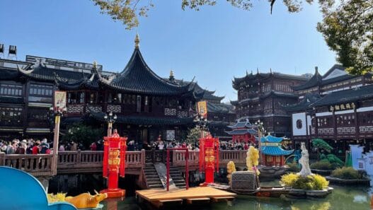 Traditionele Chinese architectuur omringt een vijver met rode lantaarns, beelden en een houten brug vol mensen onder een helderblauwe lucht. Hierdoor is uw tuin een bezoek waard vanwege de levendige sfeer en culturele schoonheid. -copyright-moving-jack.com