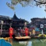 Traditional chinese architecture surrounds a pond with red lanterns, statues, and a wooden bridge crowded with people under a clear blue sky, making yu garden worth visiting for its vibrant atmosphere and cultural beauty. -copyright-moving-jack. Com