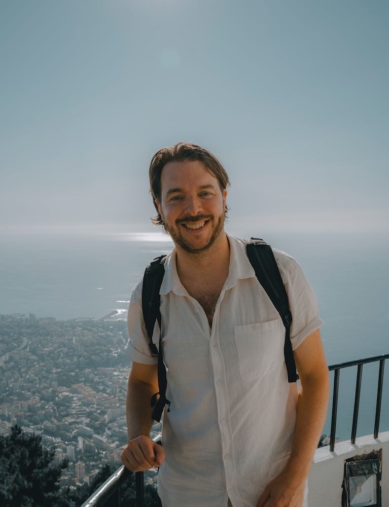 About moving jack 6 about me A man wearing a white shirt and backpack stands smiling at a scenic overlook with a city and ocean in the background under a clear sky. -copyright-moving-jack. Com