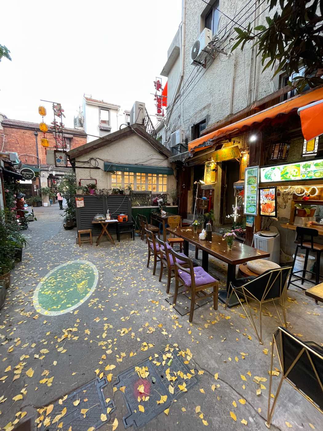 Outdoor dining area with empty tables and chairs along a narrow street in shanghai at night, surrounded by buildings and scattered yellow leaves on the ground. -copyright-moving-jack. Com