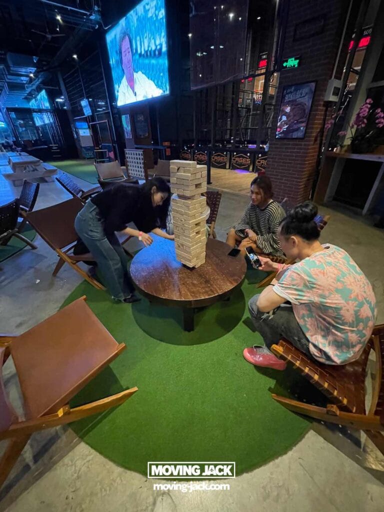 Shanghai 's nachts: beste handboek voor het nachtleven (2025) 13 Shanghai 's nachts Drie mensen zitten rond een ronde tafel en spelen binnenshuis een groot Jenga-spel. Door de ramen zijn de felle lichten van Shanghai 's nachts zichtbaar, terwijl één persoon een blok trekt en anderen aandachtig toekijken. -copyright-moving-jack.com