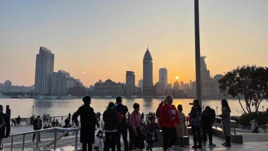 A group of people stands on a city waterfront promenade at sunset, with tall buildings and a river in the background, capturing the vibrant energy of shanghai at night. -copyright-moving-jack. Com
