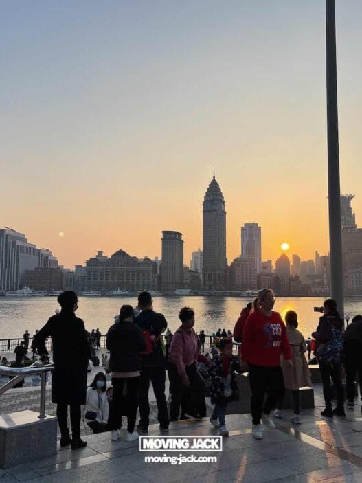 15 unique things to do in shanghai near the bund 50 renting a car in okinawa A group of people stands on a city waterfront promenade at sunset, with tall buildings and a river in the background, capturing the vibrant energy of shanghai at night. -copyright-moving-jack. Com