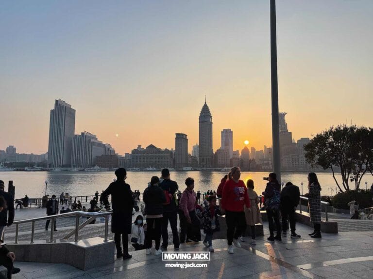 A group of people stands on a city waterfront promenade at sunset, with tall buildings and a river in the background, capturing the vibrant energy of shanghai at night. -copyright-moving-jack. Com