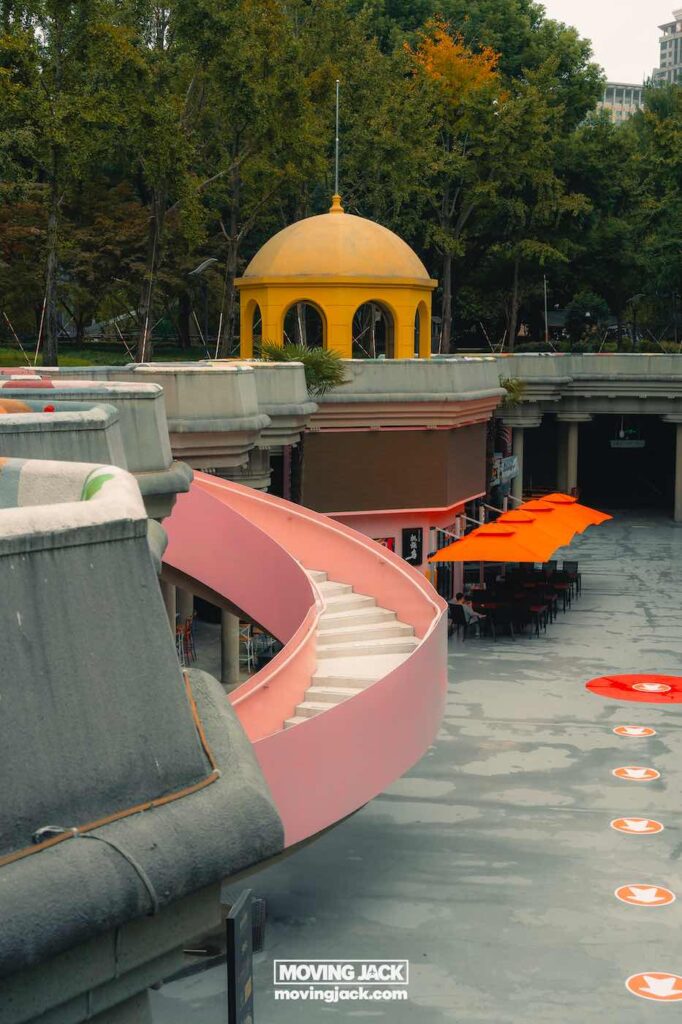 Shanghai 's nachts: beste handboek voor het nachtleven (2025) 12 Shanghai 's nachts Een gebogen roze trap leidt naar een lager gelegen buitenruimte met oranje parasols en een gele koepelstructuur, omgeven door bomen, die de levendige energie van Shanghai bij nacht vastleggen. -copyright-moving-jack.com