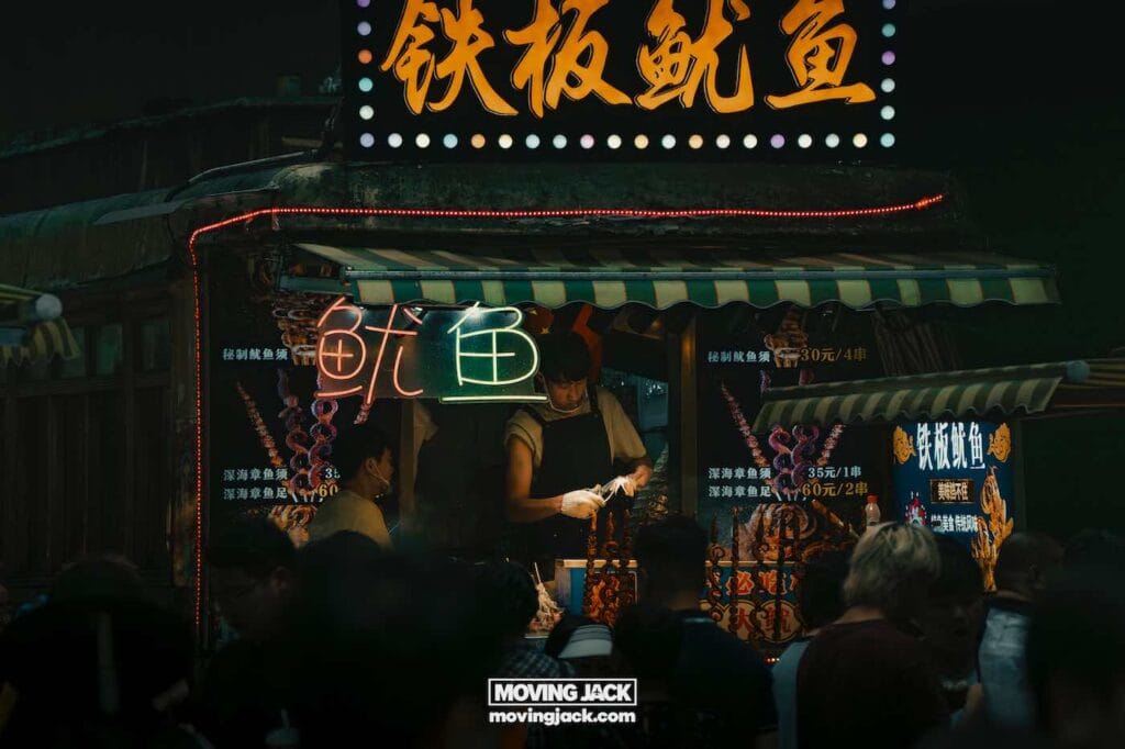 A vendor prepares food at a brightly lit street food stall with neon signs in Chinese.