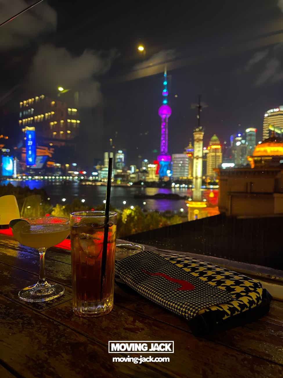 57 things to do in shanghai: the ultimate handbook 24 things to do in shanghai Two cocktails and a patterned clutch rest on a table at a shanghai rooftop bar overlooking the vibrant skyline at night, with illuminated skyscrapers and the oriental pearl tower creating the perfect backdrop for luxury things to do in shanghai.
