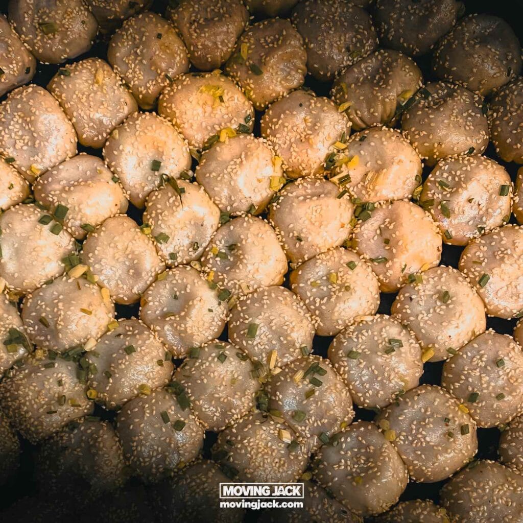 A close-up view of several pan-fried Xiaolongbao dumplings topped with sesame seeds and chopped green onions, arranged closely together.