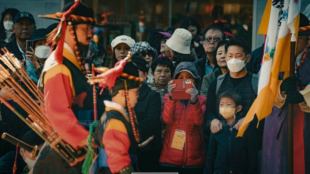 A crowd enjoys a traditional cultural parade—one of the free things to do in seoul—with two people in colorful historical costumes. Some spectators wear masks, while another records the vibrant scene with a red phone. -copyright-moving-jack. Com